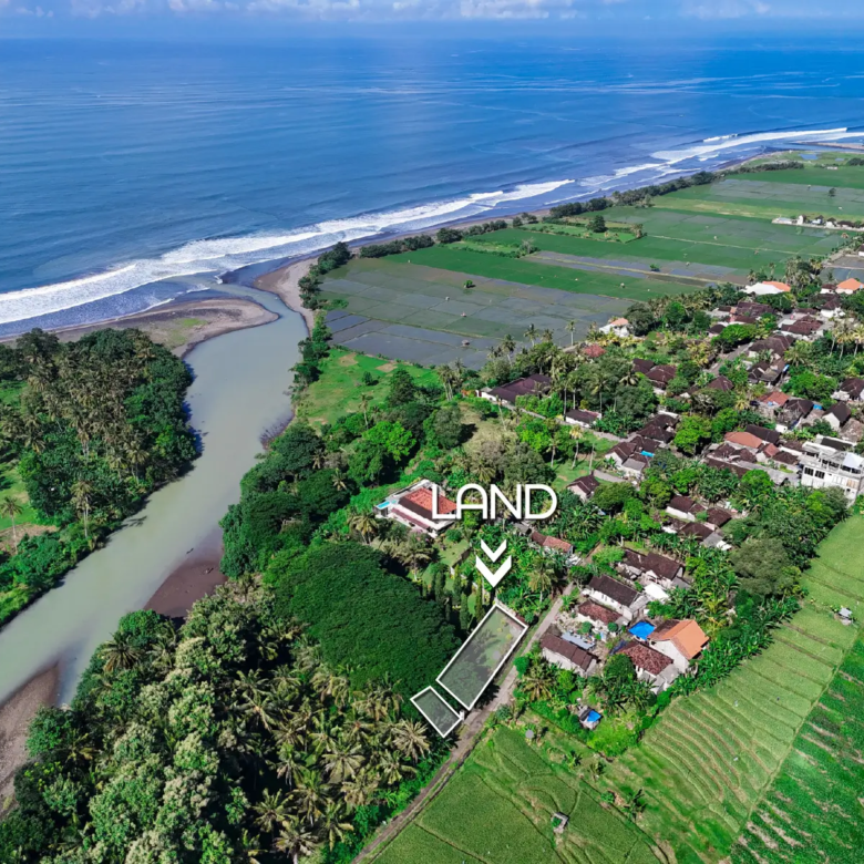 Drone view of land near river mouth in West Bali, surrounded by rice fields and village area with ocean in the background.