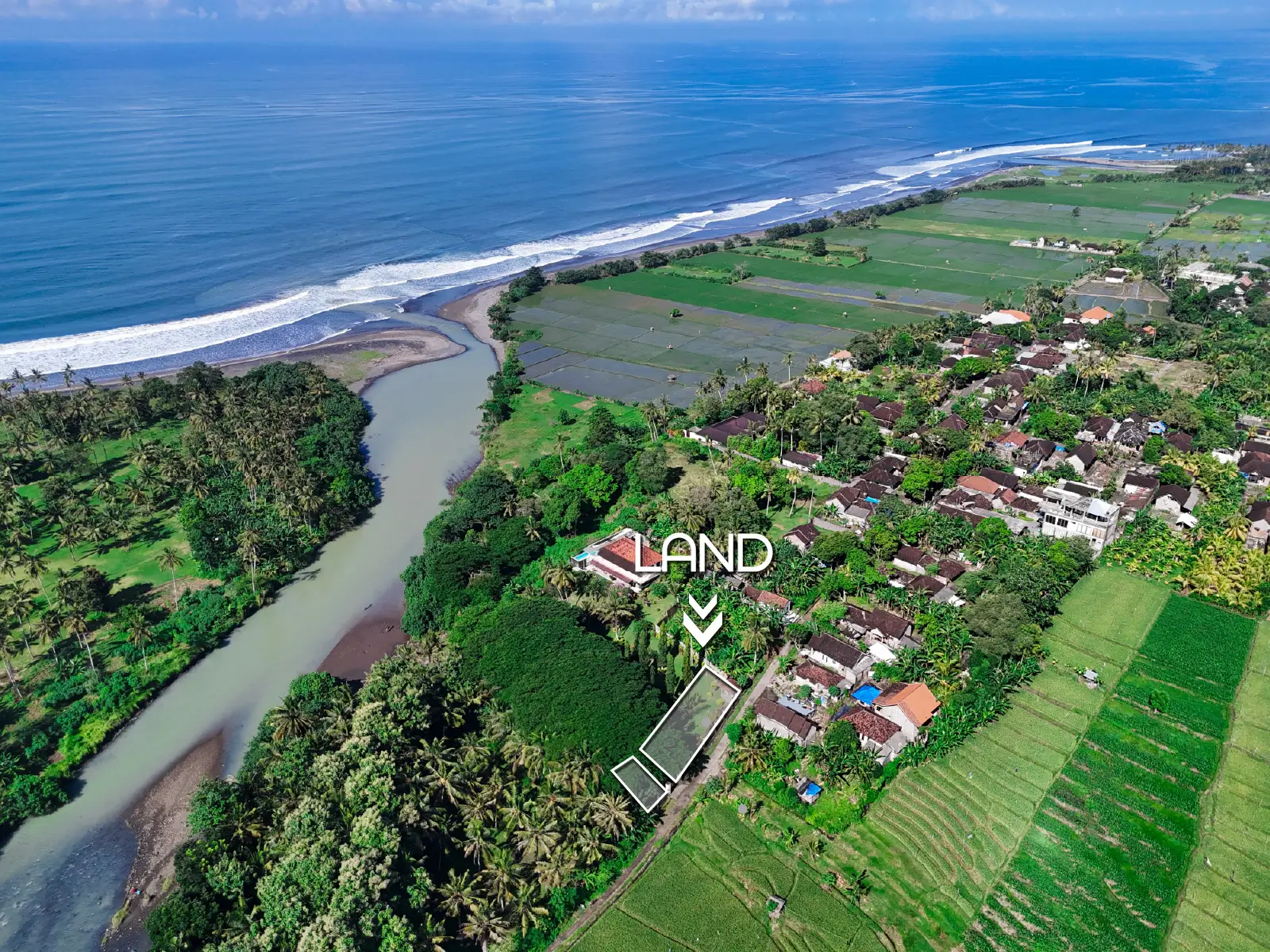 Drone view of land near river mouth in West Bali, surrounded by rice fields and village area with ocean in the background.
