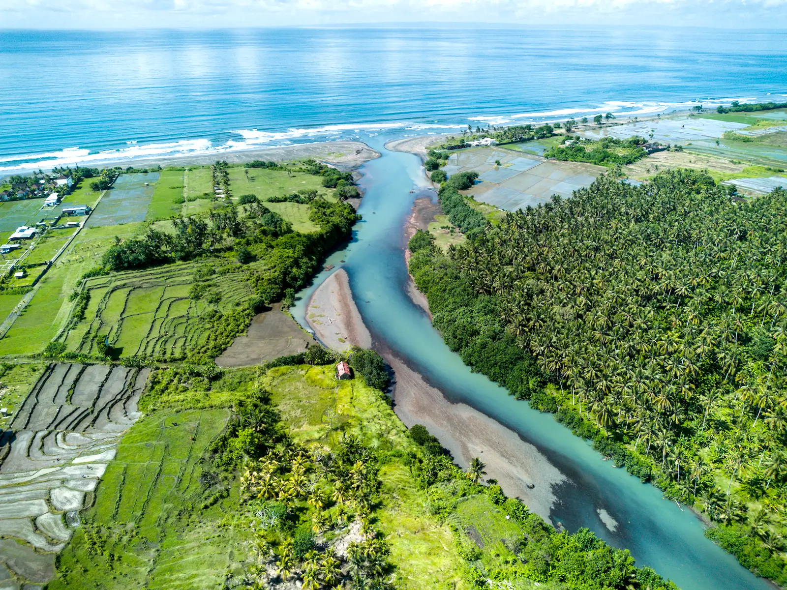 Wide aerial view of West Bali showing a river estuary meeting the ocean, surrounded by green rice terraces and tropical vegetation