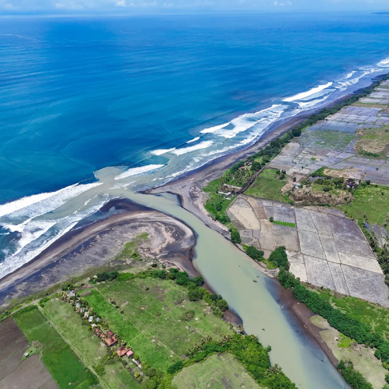 Wide aerial coastal landscape of West Bali featuring ocean waves, river estuary, and expansive rice fields stretching along the shoreline