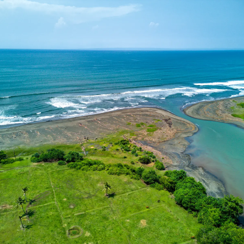 Scenic aerial view of West Bali beachfront land where the river meets the ocean, showing wide sandy coastline, blue waves, and lush green landscape.