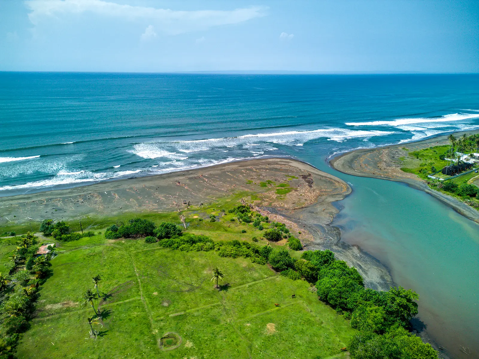 Scenic aerial view of West Bali beachfront land where the river meets the ocean, showing wide sandy coastline, blue waves, and lush green landscape.