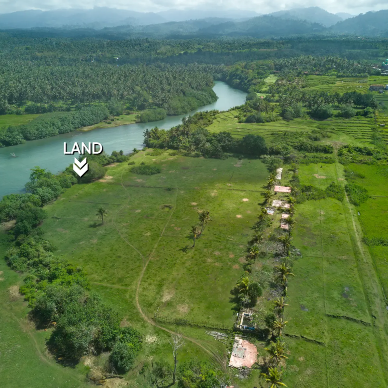 Aerial photo of riverfront land for sale in West Bali, featuring wide green fields, coconut trees, mountain backdrop, and direct access to the river.