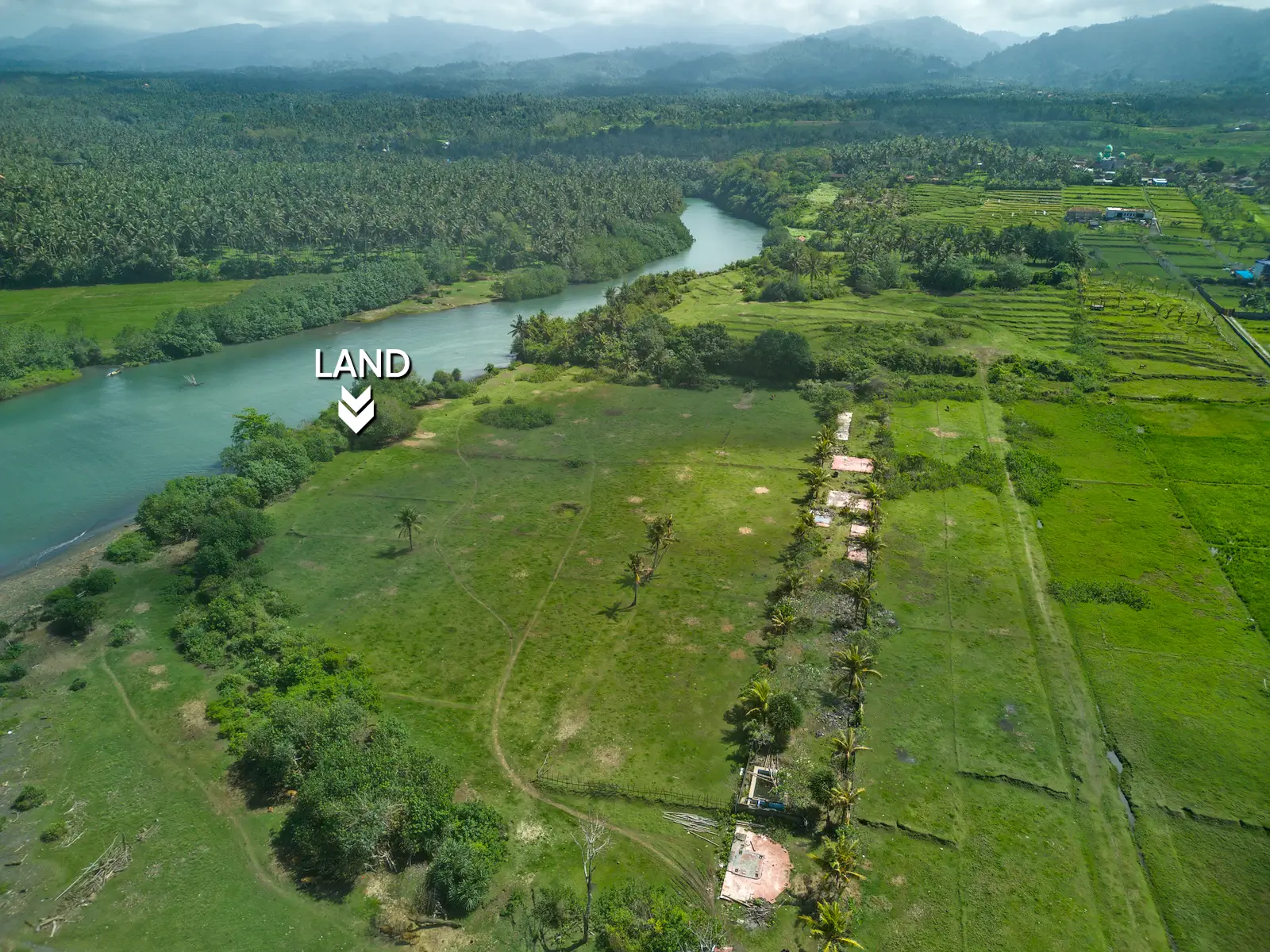 Aerial photo of riverfront land for sale in West Bali, featuring wide green fields, coconut trees, mountain backdrop, and direct access to the river.