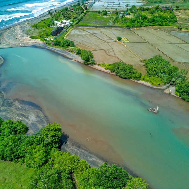 Scenic aerial view of the river lagoon meeting the ocean in West Bali, featuring clear turquoise water, coastal landscape, and beachfront land in a natural setting.