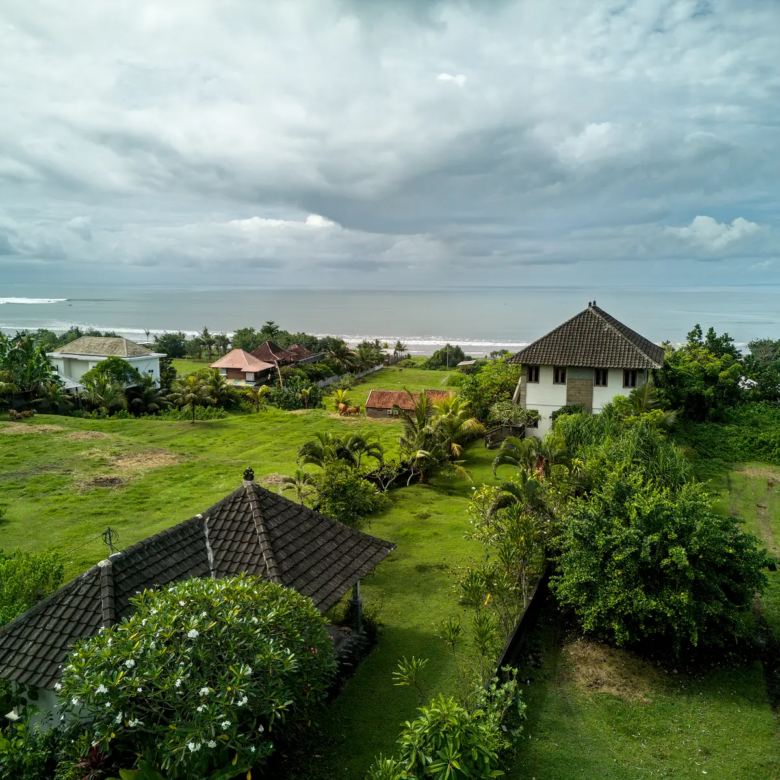 Ground-level view from a land plot in West Bali, featuring surrounding villas, tropical gardens, and ocean waves visible on the horizon