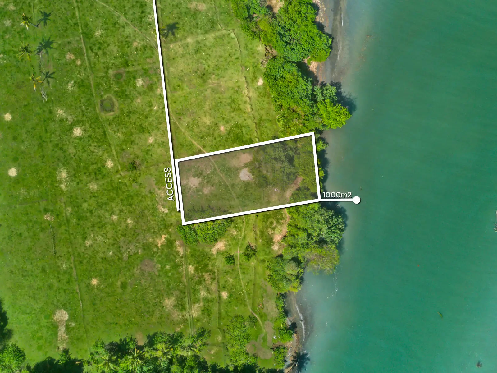 Aerial view of a 1,000 m² riverfront land plot in West Bali, located steps from the beach with turquoise lagoon water, ocean waves, and open green fields.